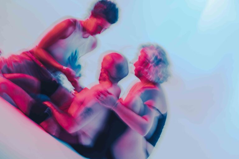 Drie mensen zitten in een wazige compositie, waarbij één persoon zich over de ander gebogen heeft, met een creatieve verlichting die een mix van roze en blauwe tinten creëert.