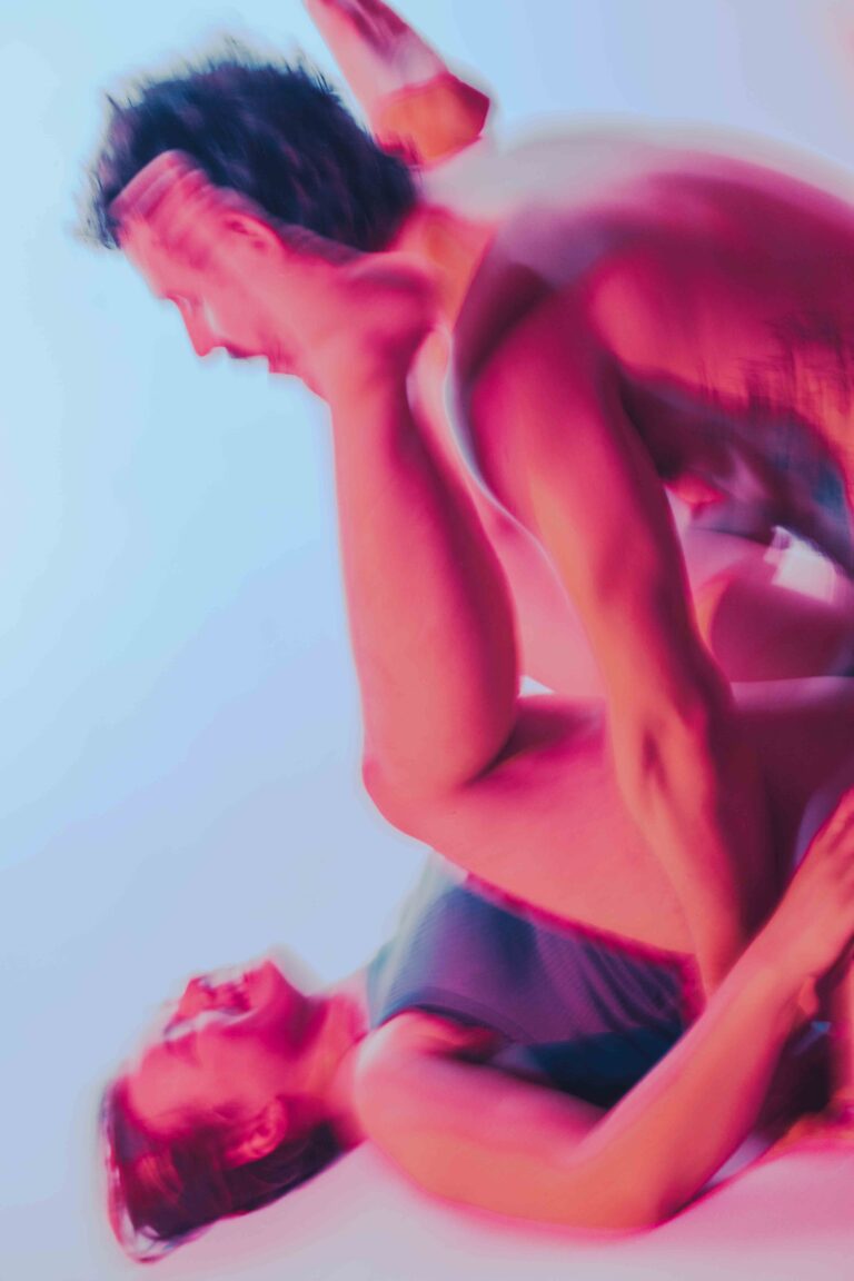 Two nude performers in an artistic pose, one person lifting or supporting another, captured in vibrant pink and purple hues against a soft blue background.