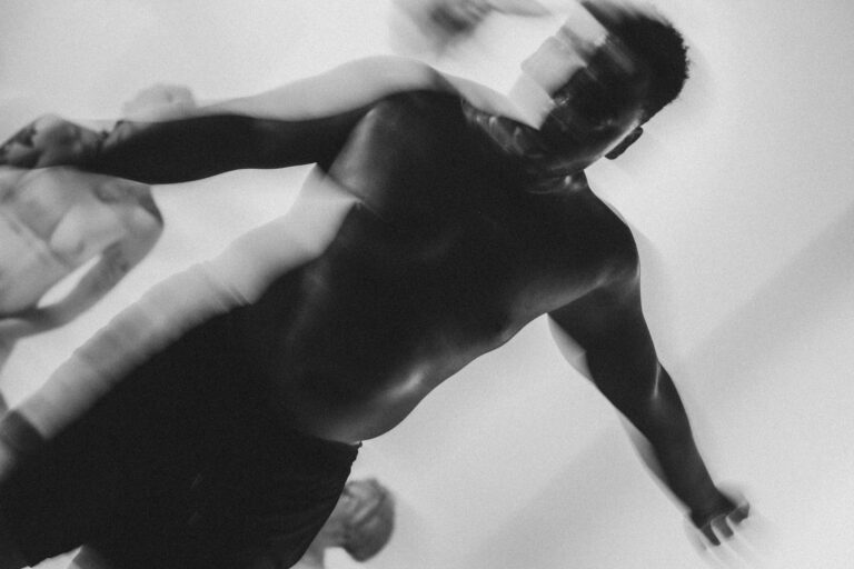 Een groep naakte mensen met Philemon Mukarno die elkaar ondersteunen in een acrobatische houding, met een persoon in het midden die door anderen wordt vastgehouden. De foto is in zwart-wit en geeft een artistieke sfeer weer, met een focus op de menselijke vormen en bewegingen.