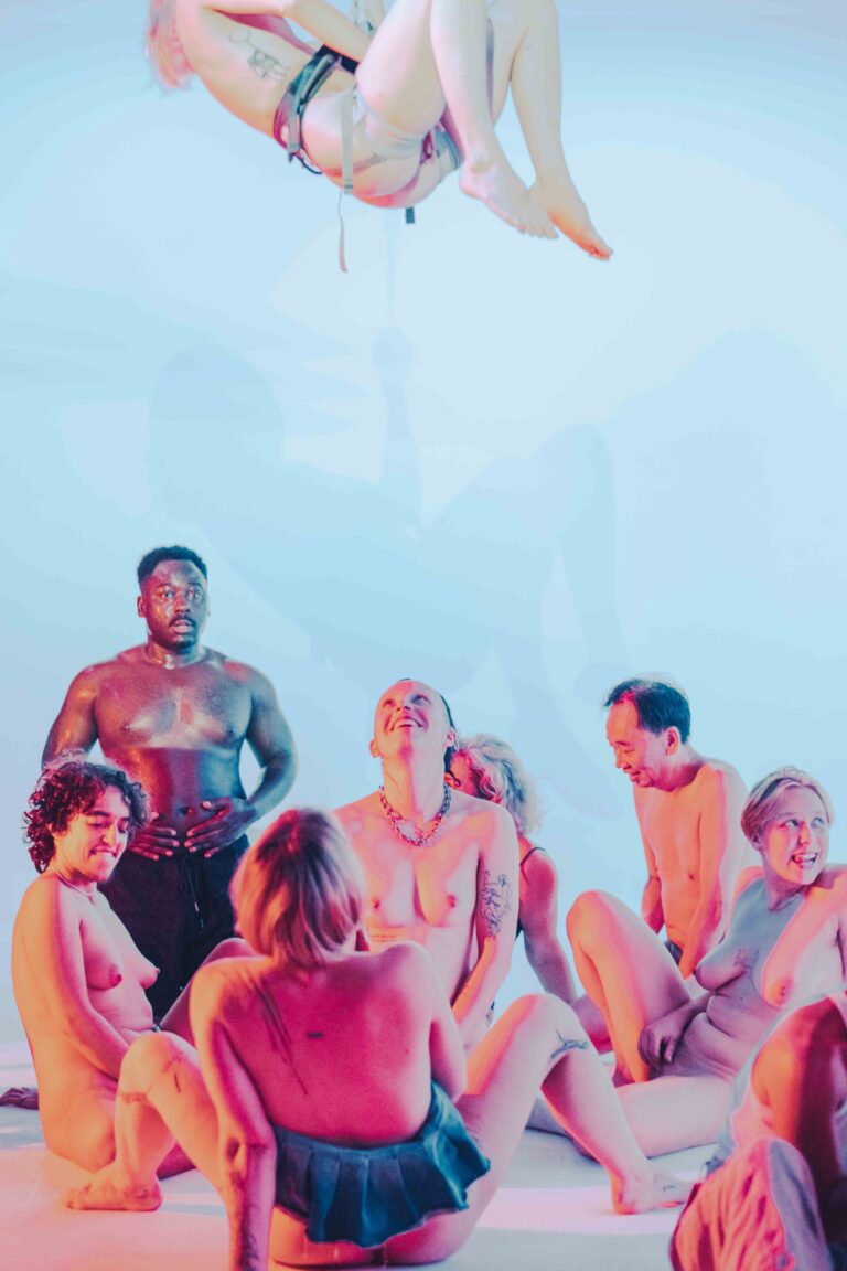 Circle of mostly nude performers sitting on floor watching aerial artist performing above them in soft blue atmosphere