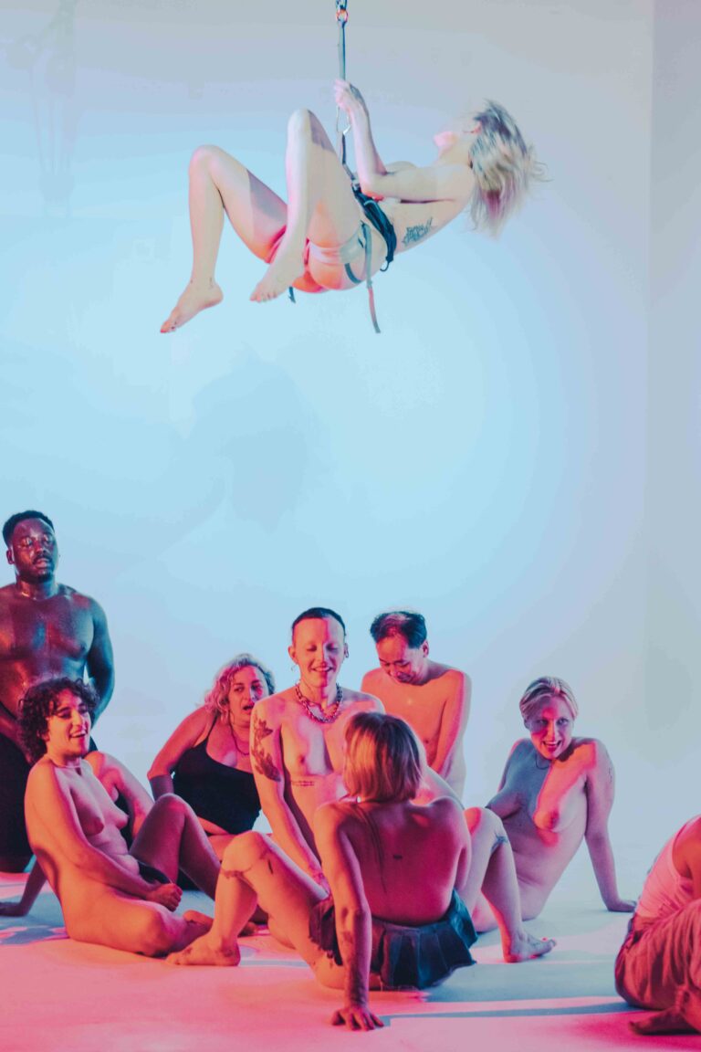 Aerial performer suspended above group of nude performers sitting in circle formation on light-colored floor
