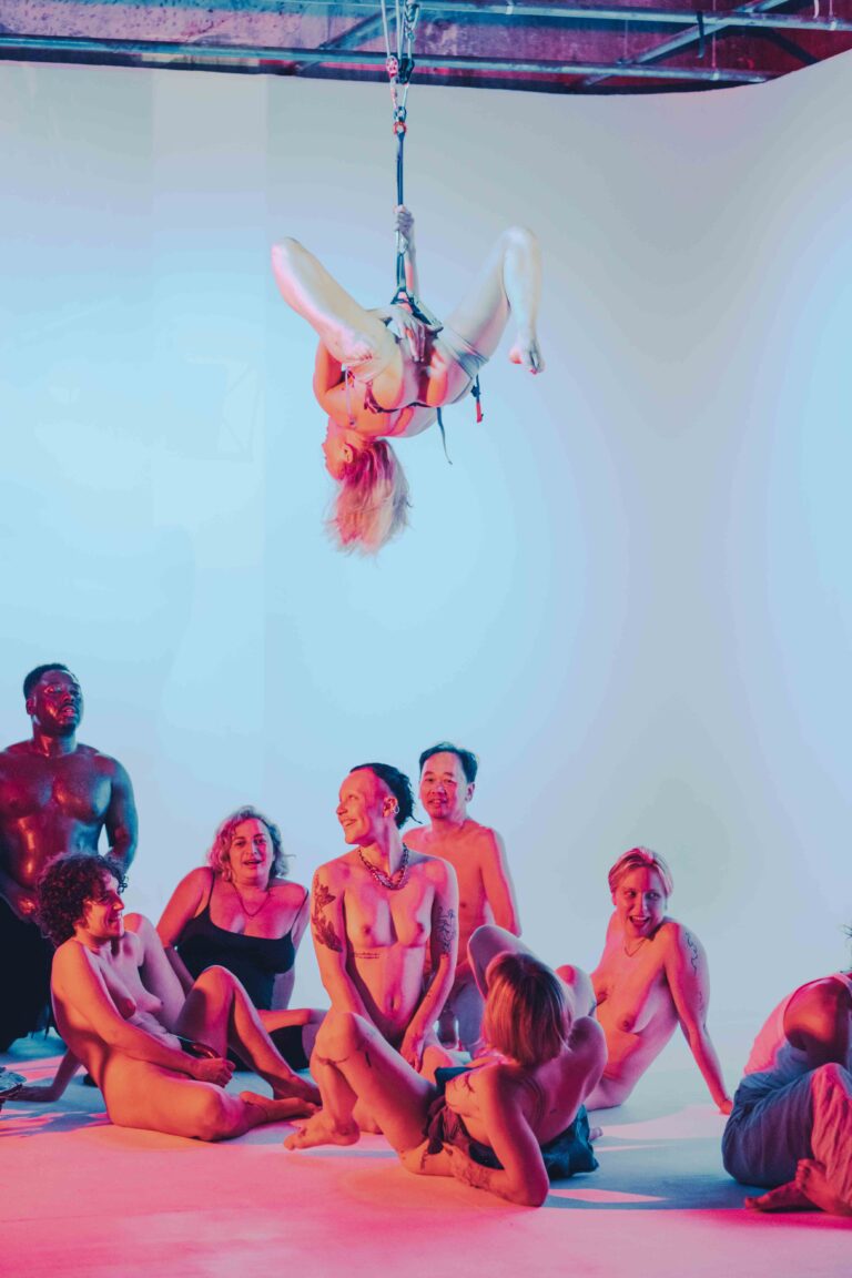 Aerial performer hanging upside down above circle of seated nude performers, industrial ceiling beams visible in pink and blue light
