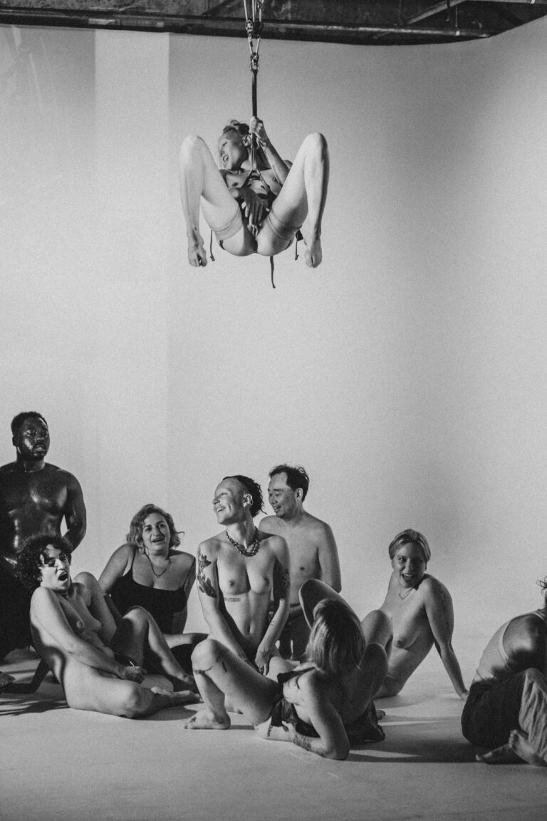 Group of nude performers sitting on the ground looking up at one person suspended upside down from chains in the center
