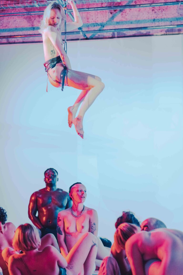 Blonde woman suspended on rope while group of nude performers sit below, industrial rafters visible above in pink lighting