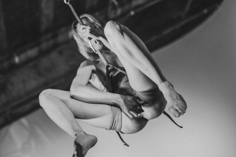 Naakte persoon hangt aan een touw in een acrobatische pose, met de benen gebogen en de armen om het touw geklemd, tegen een zachte, onduidelijke achtergrond in zwart-wit tinten.