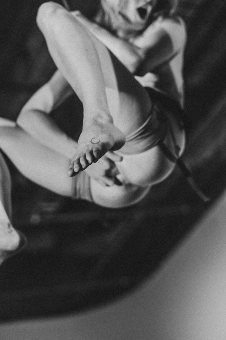 Naked woman in an acrobatic pose suspended from chains looking down at the camera