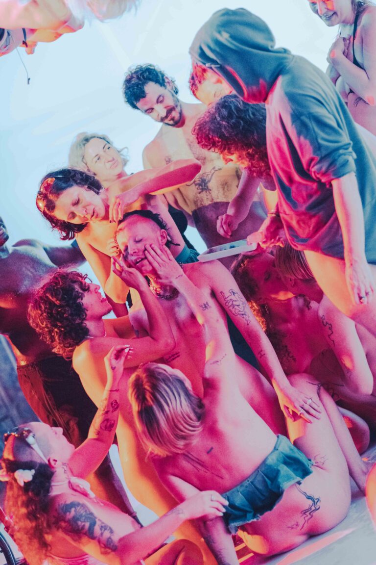 Group of nude performers with Philemon Mukarno on stage, captured from above in vibrant pink and blue lighting