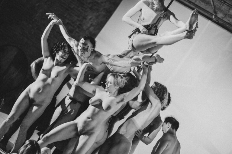 Een groep naakte mensen met Philemon Mukarno die elkaar ondersteunen in een acrobatische houding, met een persoon in het midden die door anderen wordt vastgehouden. De foto is in zwart-wit en geeft een artistieke sfeer weer, met een focus op de menselijke vormen en bewegingen.