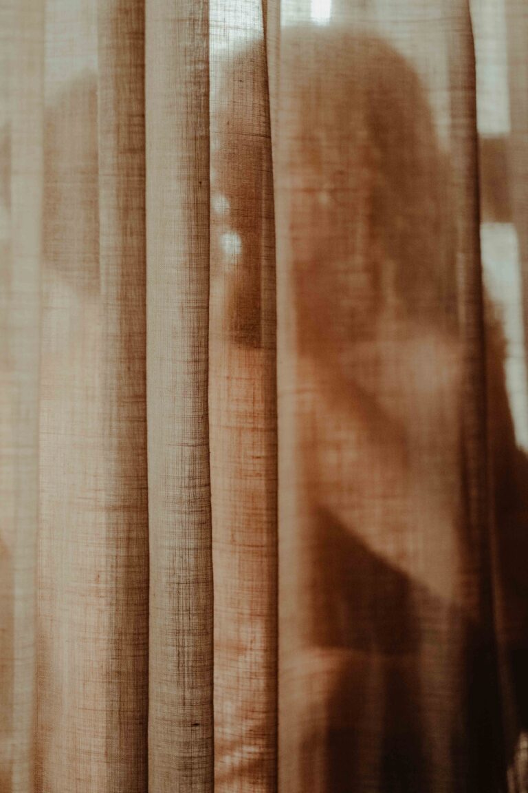 Blurred silhouette of a nude person behind a sheer curtain in soft natural lighting