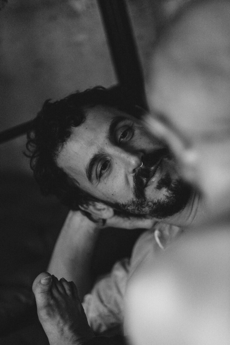 Close-up black and white image of a shirtless man with curly hair and beard looking sideways at camera