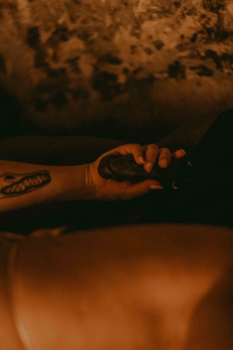 A tattooed hand holds a small furry object against a nude body in warm, atmospheric lighting