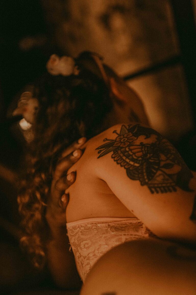 Close-up of a topless female figure with a large tattoo on the arm, wearing lace underwear in warm, moody lighting