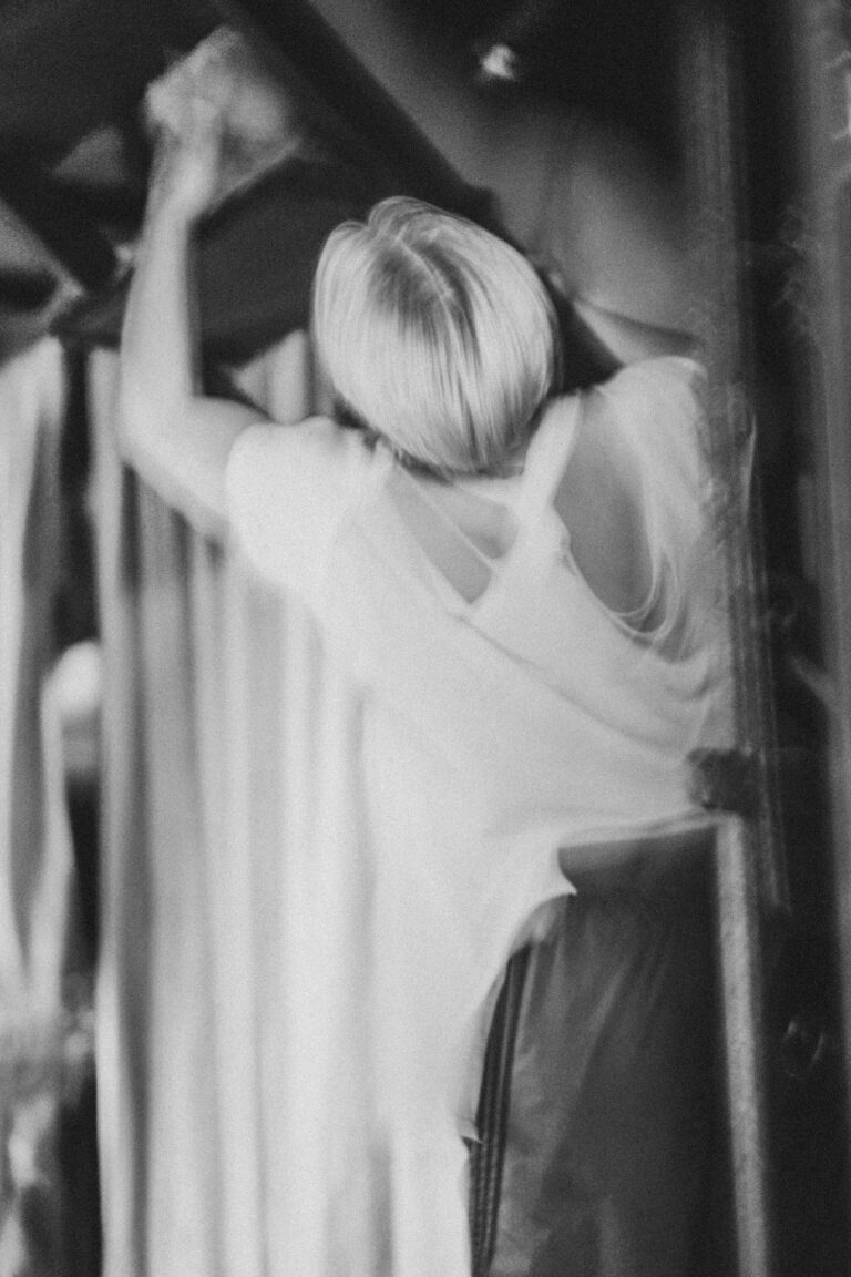 Naked person with short blonde hair standing behind white curtains reaching upward