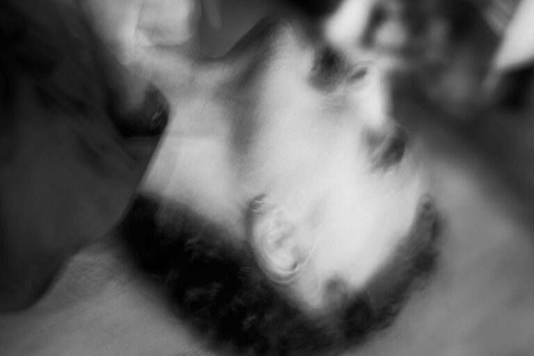 Een close-up van een man met een lichte baard en een ring in zijn oor, terwijl een hand met een latexhandschoen zijn kin vasthoudt. De achtergrond is vaag en donker, wat een dramatische sfeer creëert.