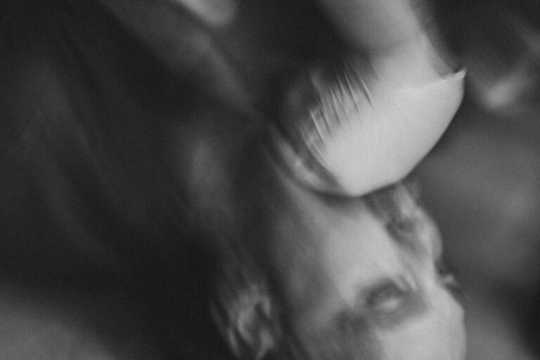 Een close-up van een man met een lichte baard en een ring in zijn oor, terwijl een hand met een latexhandschoen zijn kin vasthoudt. De achtergrond is vaag en donker, wat een dramatische sfeer creëert.