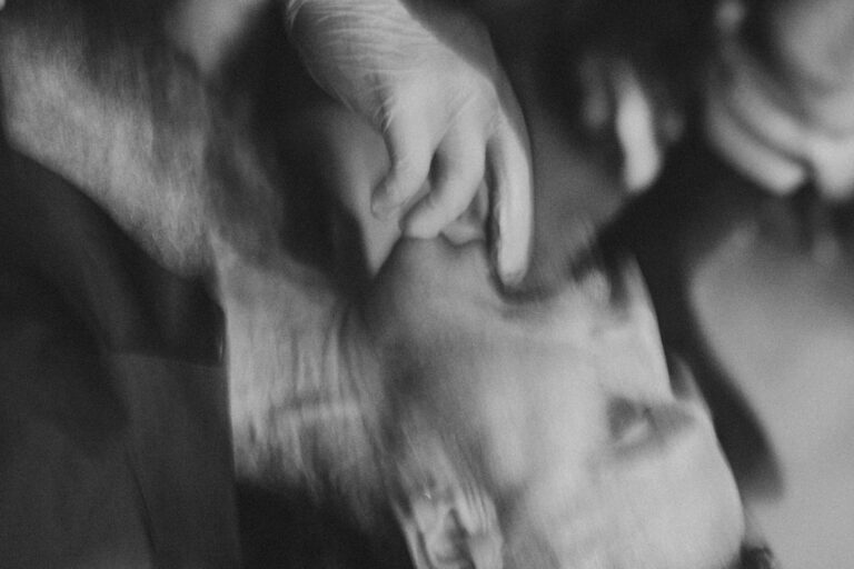 Een close-up van een man met een lichte baard en een ring in zijn oor, terwijl een hand met een latexhandschoen zijn kin vasthoudt. De achtergrond is vaag en donker, wat een dramatische sfeer creëert.