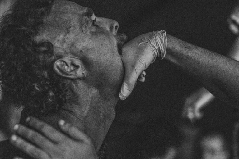 Een close-up van een man met een lichte baard en een ring in zijn oor, terwijl een hand met een latexhandschoen zijn kin vasthoudt. De achtergrond is vaag en donker, wat een dramatische sfeer creëert.