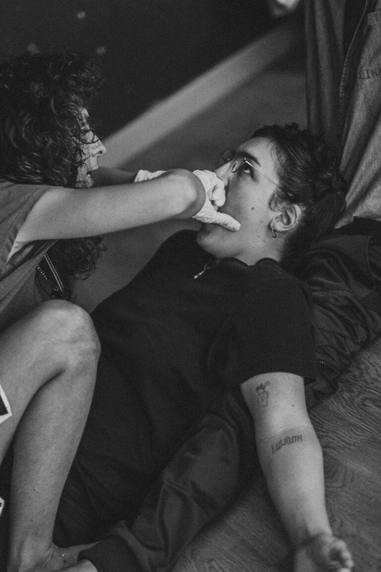 Naked person with short blonde hair reclining on dark fabric with hand raised to face