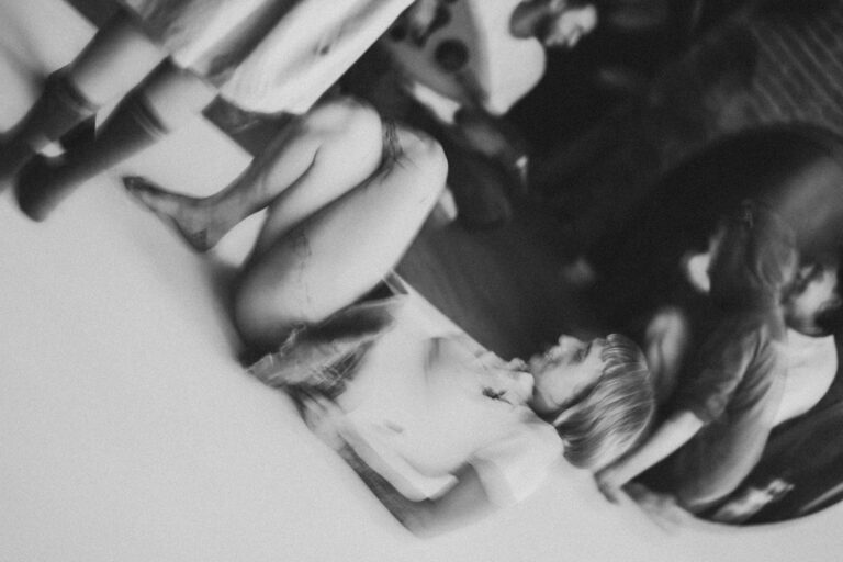 Een zwart-wit afbeelding van een vrouwelijke persoon die op de grond ligt, omringd door andere mensen die zich bezig houden met verschillende activiteiten. De achtergrond is onscherp en het beeld heeft een dromerige sfeer.