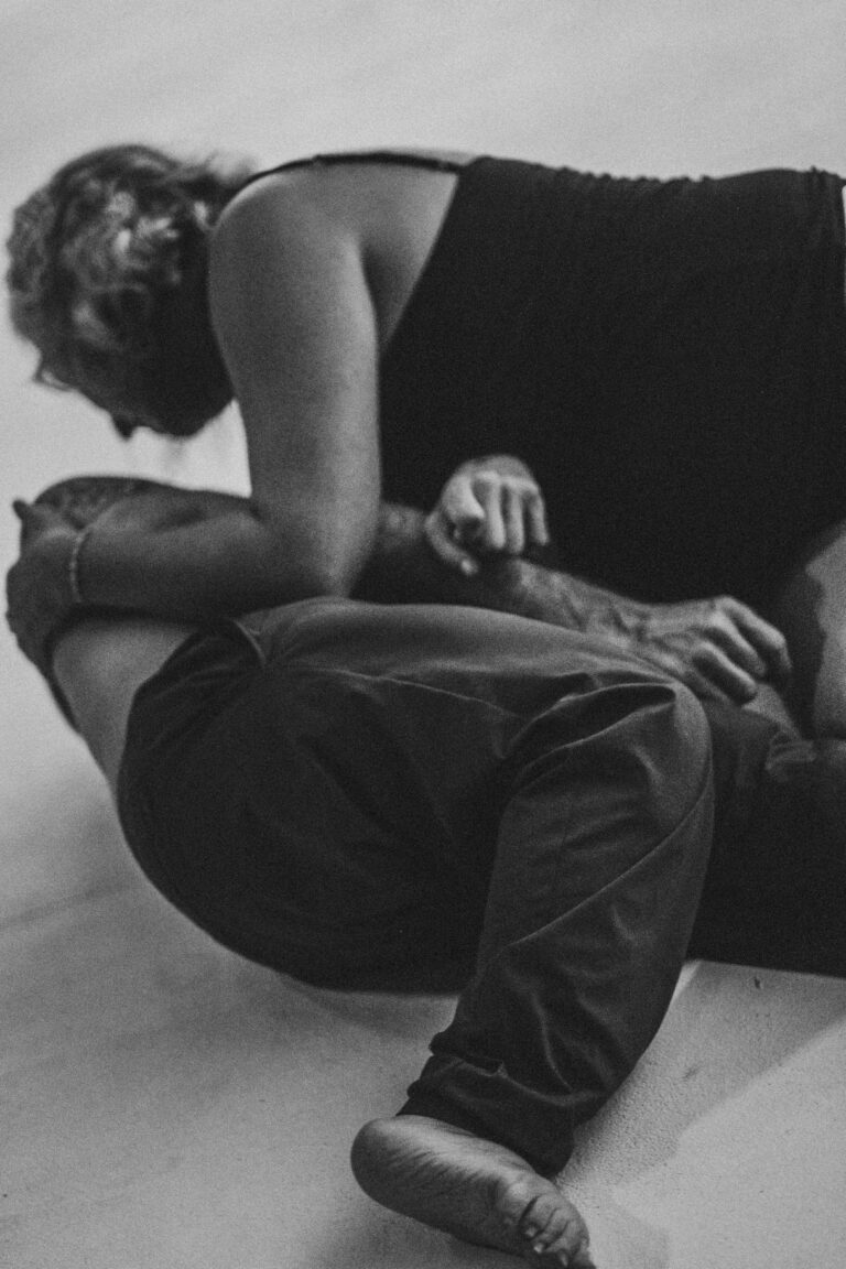 Two women, one with short curly hair topless crouching on the ground, one fully clothed sitting beside her