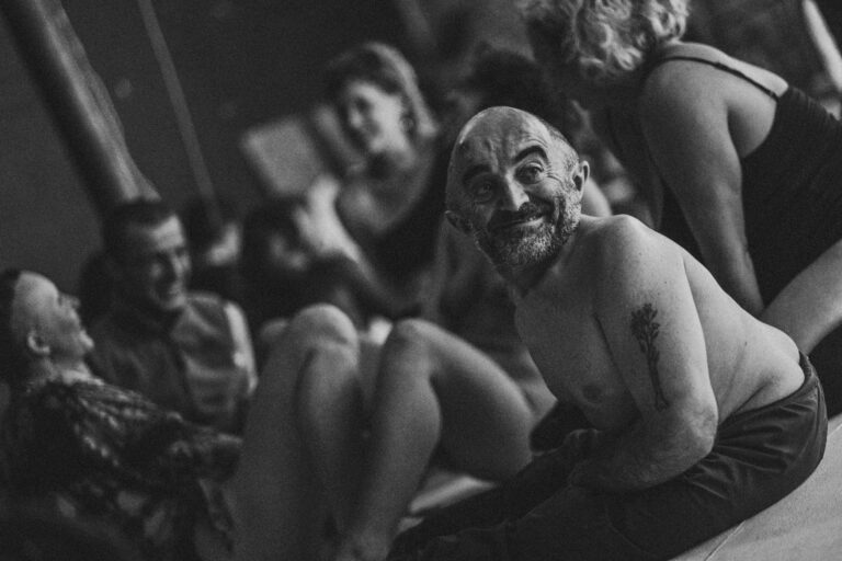 Een zwart-wit afbeelding van een vrouw met halflang haar die een man met een kaal hoofd en een baard aanraakt, terwijl hij zijn ogen gesloten heeft en een neutrale uitdrukking op zijn gezicht vertoont. De vrouw is deels uit beeld en draagt een zwart kledingstuk, terwijl de man geen bovenkleding draagt. De setting lijkt intiem en dicht bij de camera te zijn genomen.