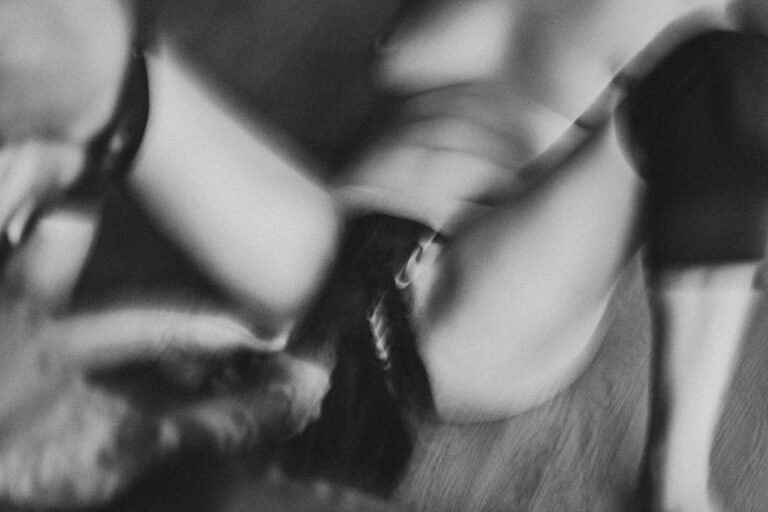 Een zwart-wit afbeelding van een vrouw met halflang haar die een man met een kaal hoofd en een baard aanraakt, terwijl hij zijn ogen gesloten heeft en een neutrale uitdrukking op zijn gezicht vertoont. De vrouw is deels uit beeld en draagt een zwart kledingstuk, terwijl de man geen bovenkleding draagt. De setting lijkt intiem en dicht bij de camera te zijn genomen.