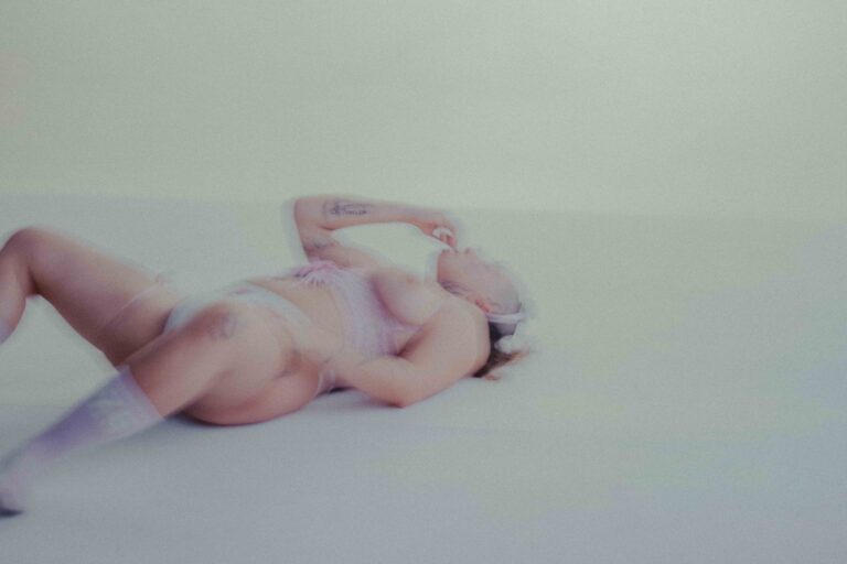 Een vrouw in lingerie ligt op de grond met een zachte, wazige achtergrond. Ze heeft tatoeages en draagt een haarband.