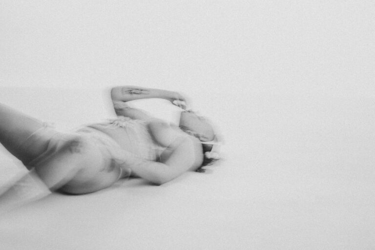 Een vrouw ligt op een lichte achtergrond, gekleed in lingerie, met een hand achter haar hoofd en de andere hand naar haar mond reikend. Haar houding en de beweging van de camera creëren een dromerige, ethereal sfeer.