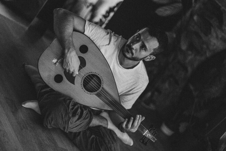 A man sits on the floor and plays a traditional lute, dressed in a white T-shirt and loose pants. The environment is dark and atmospheric, with vague, artistic backgrounds.