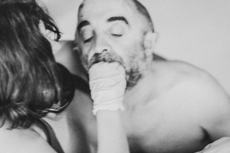 A black-and-white image of a woman with long hair touching a bald man with a beard, while he has his eyes closed and displays a neutral expression on his face. The woman is partly visible and wearing a black piece of clothing, while the man is not wearing an upper garment. The setting looks intimate and close to the camera.