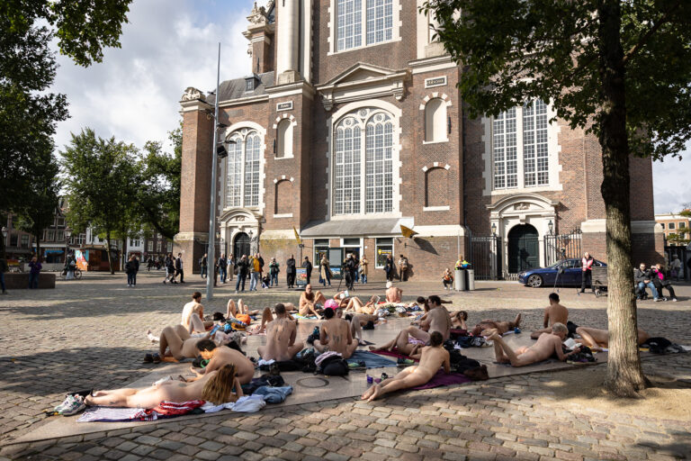 Een groep naakte mensen staat in een ruimte, terwijl anderen in groepjes op de vloer liggen. De staande personen houden rode boekjes vast en poseren of presenteren iets, terwijl de anderen zich bezig houden met verschillende activiteiten zoals lezen of ontspannen.