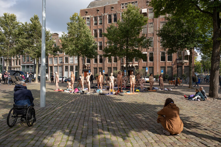 Een groep mensen in een stadspark poseert naakt op pleinen en handdoeken, terwijl enkele toeschouwers, waaronder een man in een rolstoel, zich op de achtergrond bevinden.