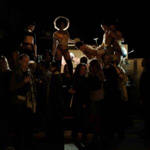 A group of people in various outfits stands in a dimly lit space, with performers in revealing costumes on a platform behind them.