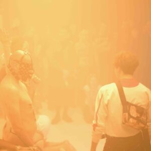 A partially clad performer with a mask stands in a foggy space, while an audience watches, appearing blurred in the background.